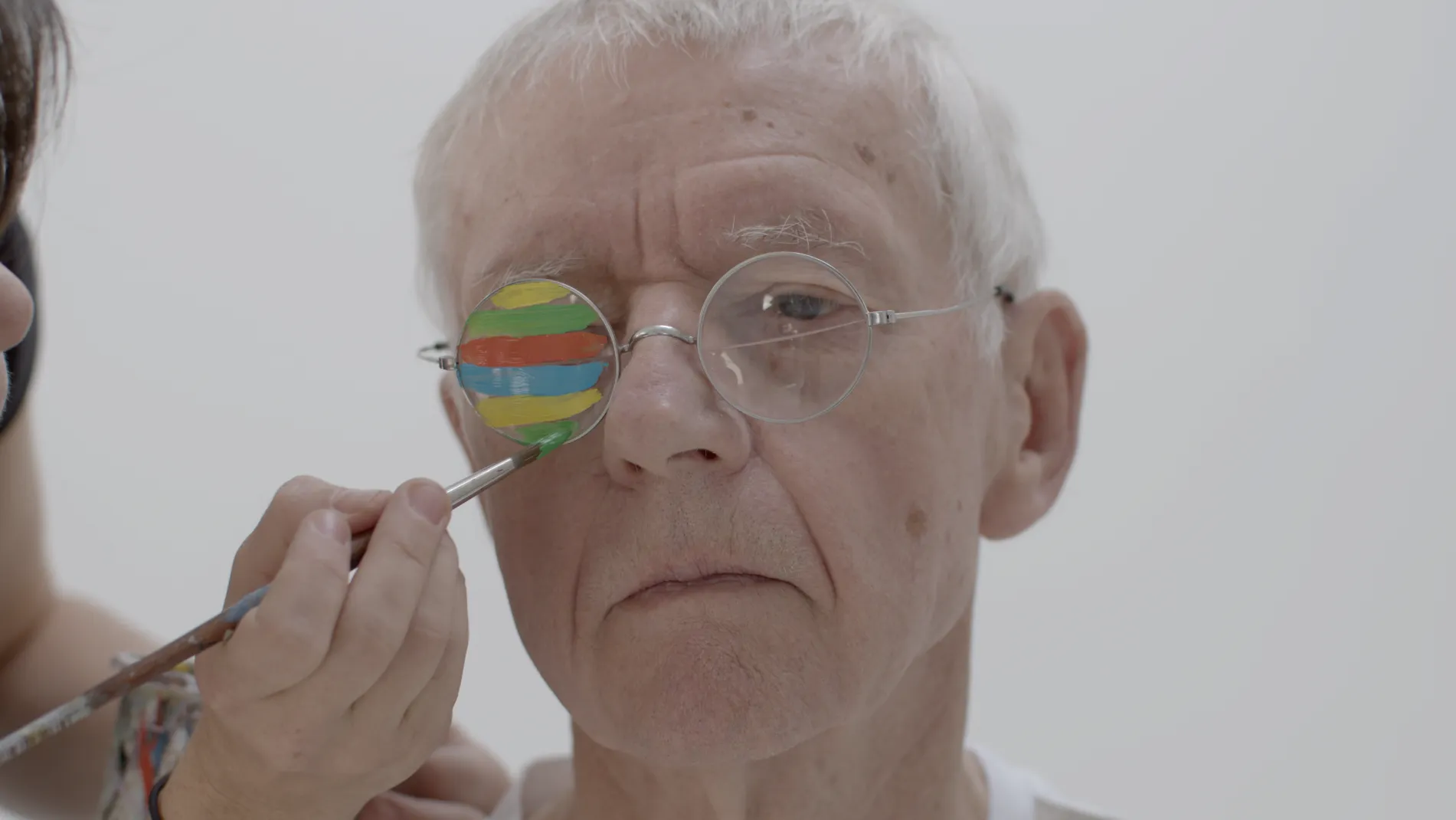Gros plan sur le visage de Daniel Authouart, peintre âgé aux cheveux blancs, un pinceau peignant en couleurs vives le verre de ses lunettes