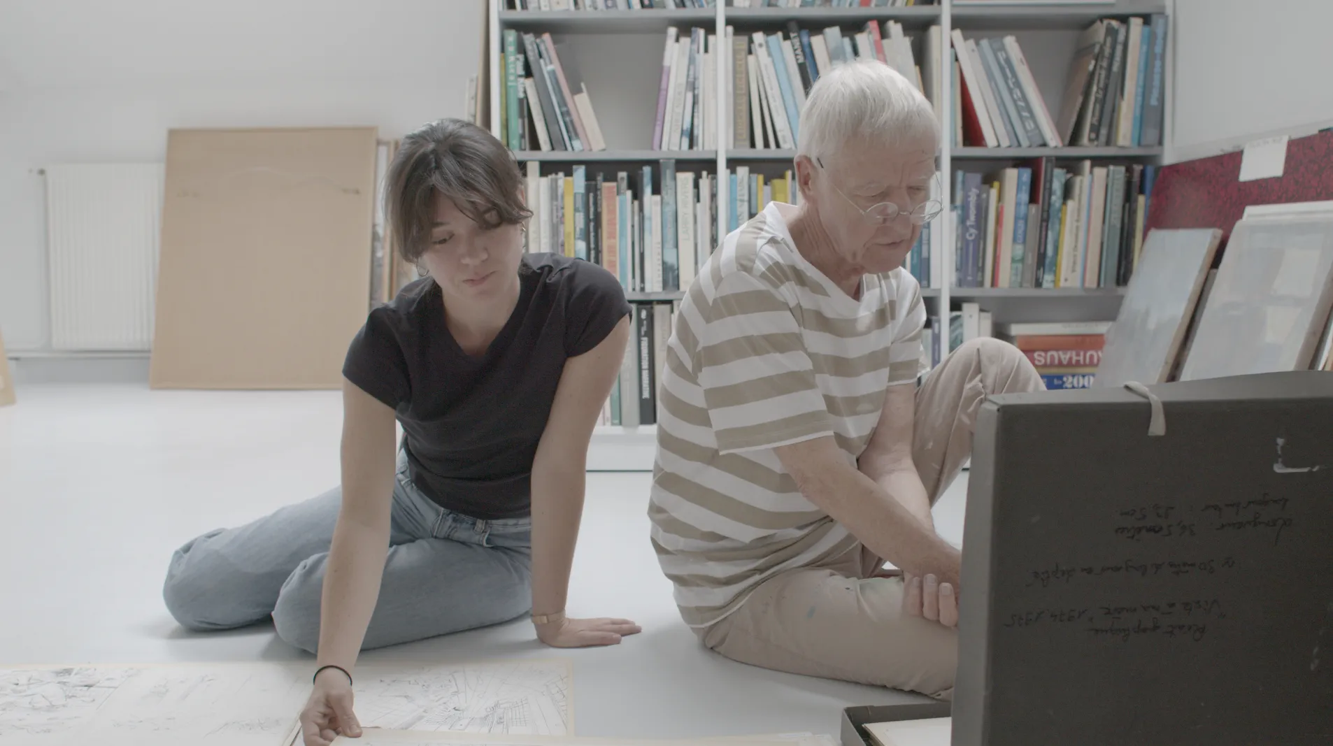 Camille Authouart et son père assis au sol, penchés sur des dessins et documents étalés devant eux, bibliothèque en arrière-plan