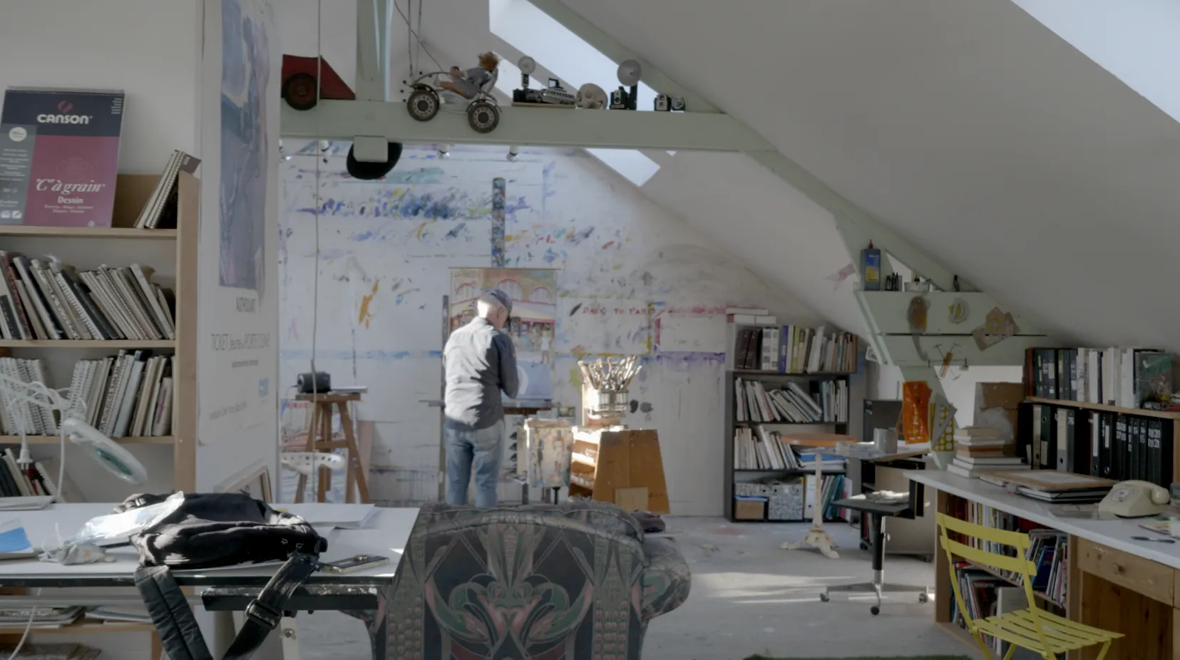 Vue d'ensemble de l'atelier de Daniel Authouart, peintre debout devant son chevalet, pièce mansardée encombrée de livres, esquisses et matériel artistique